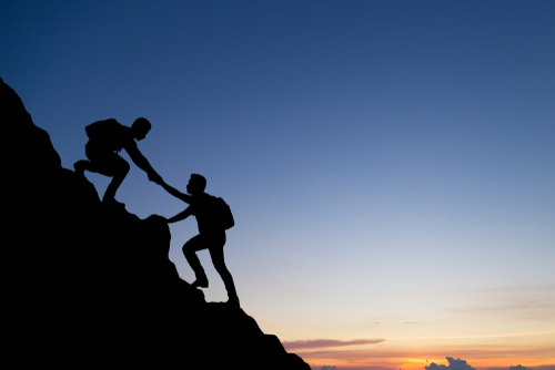 Silhouette of people climbing a mountain. One person is reaching out to help the other climb up.